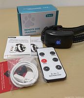 Photo showing black dog training collar with LED display, remote control, USB cable, instruction manual, and packaging