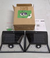 Two black solar motion sensor lights side by side with solar panel tops, LED lights below, motion sensors in middle, wall anchors, screws, and instruction manual on white surface with brown cardboard box behind