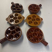 Top-down and angled view of six ceramic escargot dishes arranged on a flat surface. Three lighter brown dishes with dual side handles, three darker brown dishes with single handles. Each dish has 6 circular indentations for holding escargot.