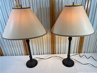 Both matching ornate table lamps with shades side by side on white surface in front of cream panel backdrop.
