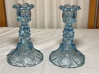 Pair of light aqua blue pressed glass candlestick holders displayed side by side on white cloth with wooden chair background