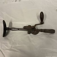 Full view of the antique breast chest drill showing the curved breast plate, crank handle, and wooden grip at the drill end on a crumpled white background.