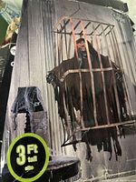 Full view of the 3-foot tall Grim Reaper behind cage Halloween decoration, shown with Halloween-themed backdrop including window and spiderweb-covered lamp.