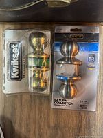 Two door knob sets in original packaging: Kwikset gold-tone set and Defiant Saturn Collection stainless steel set.