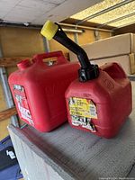 Two red plastic gas cans sitting side by side, showing handle, caps, and spout on one can.
