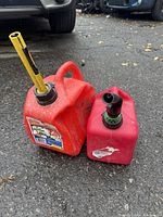 Two red plastic fuel/gas cans placed on ground in an outdoor setting with pavement and vehicles in background. Larger can has yellow and black spout with a measurement gauge, smaller can has black and green spout attachment.