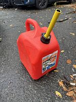 Full view of the red 5 gallon gas can with yellow spout sitting on ground outdoors.