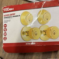 Closed packaging showing product branding and contents: Hyper Tough keyed entry doorknobs and deadbolts, polished brass finish, 2 pack.