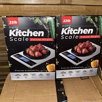 Two boxed digital kitchen scales with packaging illustrating product features and usage with strawberries on scale platform.