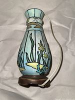 Full view of the ceramic table lamp showing the fish and plant designs, blue background, and wooden base with the electrical cord visible.