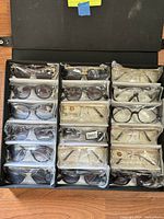 Overhead view of 18 pairs of mixed designer sunglasses in clear plastic bags inside a black box, showing variety of frame styles and colors.