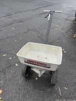Side and top perspective of the Craftsman seeder spreader's hopper, wheels, and handle on pavement.