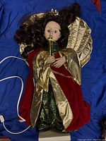 Front view of vintage Christmas doll with curly dark hair, dressed in layered red and gold gown holding a gold object. Visible electric cord on the side.