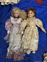 Full view of two vintage porcelain dolls in Victorian-style dresses lying on a blue fabric background