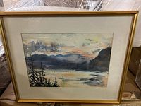 Framed watercolour landscape painting showing mountains, trees, and water with soft cloudy sky and pastel tones.