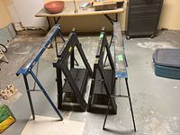 Four saw horses arranged on a concrete floor: two blue metal Mastercraft saw horses and two black Stanley plastic saw horses.
