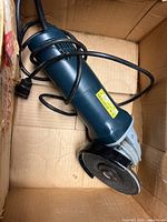 Angle grinder tool in cardboard box, showing body, power cord, and abrasive disc attached.
