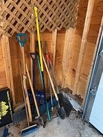 Overall view of tools leaning against wooden lattice wall in shed corner, showing multiple shovels, broom, garden hoe, and ice breaker.