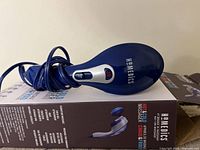 Blue and gray handheld HoMedics massager showing device logo and control panel with hot and cold functions, placed on product box.