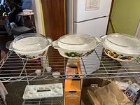 Three white ceramic casserole dishes with colorful Winter Fruits patterns and clear glass lids displayed on metal rack.