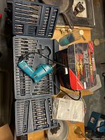 Full view of the Makita blue corded drill lying in a case with various drill bits set and a box showing the packaging.
