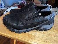 Pair of black Merrell Polarand Rove Moc women's slip-on shoes on a wooden surface, side view showing the Merrell logo on one shoe.