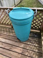 Full view of teal Rubbermaid garbage bin standing on wooden deck outdoors showing lid and body with side handles.