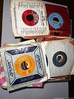 Photo showing several 45 RPM records in original paper sleeves including branded sleeves from London and Polydor. Mix of label colors seen, some records stacked in box.