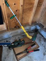 Full view of lot contents including Weed Eater string trimmer, Black & Decker hedge trimmer, manual hedge shears, and extension cord on box in wooden shed.