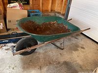 Side view of green steel wheelbarrow showing the rusted metal basin and wooden handles resting on garage floor.