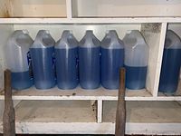 Seven semi-translucent plastic jugs of blue windshield washer fluid lined up on a shelf, showing two partially used bottles.