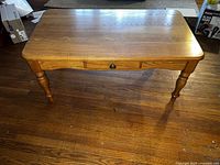 Wood coffee table showing entire top and four turned legs, displaying natural wood grain, centered drawer with round knob.