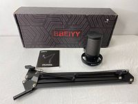 Photo showing the BBEIYY USB gaming microphone disassembled with adjustable arm stand, shock mount, user manual, and branded packaging box on white surface.