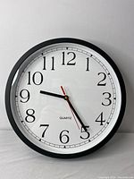 Front view of black wall clock with white clock face and black Arabic numerals, showing three clock hands and quartz label.