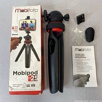 Mobifoto Mobipod TT25 mini tripod with foldable legs, ball head, Bluetooth remote shutter, phone holder clamp, and instruction sheet, laid out.