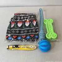 Overview of all included items on a gray surface: folded holiday dog sweater in black with Santa faces, heart, snowflakes and gifts pattern; blue ball with rope chew toy; green bone-shaped chew toy; yellow reflective collar labeled size small.