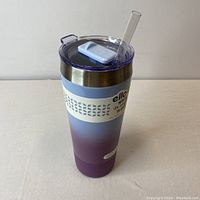 Front view showing gradient colored insulated travel mug with clear lid and light blue silicone grip band with branding.