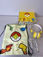 Photo showing yellow wired kids headphones with cartoon print, a matching drawstring bag, and a decorated box with colorful cartoon designs