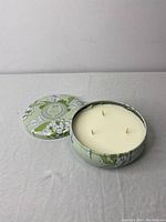 Image of the candle with lid removed showing three wicks and white wax inside a green floral printed metal container.