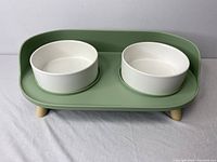 Front view of the minimalist pet feeding station showing two white bowls set in a sage green elevated tray with wooden legs.