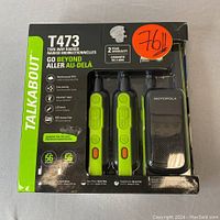 Front view of boxed Motorola T473 3-set two-way radios. Box shows two radios and charging tray visible through plastic window. Green and black color scheme with product features listed on box front.