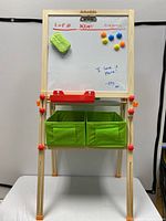 Front view of the easel featuring the dry-erase whiteboard side with attached eraser, colored magnets, red tray, and green storage bins below.