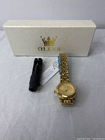 Photo of the gold tone Olevs wristwatch, box, and a black plastic tool likely for adjusting the band or battery.
