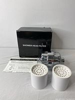 Photo showing the chrome shower head filter unit along with two white replacement filter cartridges and the installation instructions, with the product box in the background.
