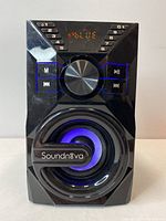 Front view of Soundnova Bluetooth speaker showing numeric keys, control buttons, large silver volume knob, and blue LED illuminated speaker cone.