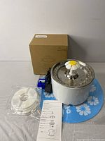Stainless steel top pet fountain with power adapter, instruction manual, spare filter, and original box on blue floral mat.