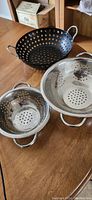 Photo showing three metal strainers on a wooden table: two stainless steel strainers and one black metal strainer, each with two handles, in good condition.