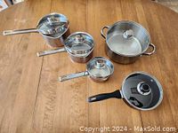 Four stainless steel pots with lids and one black aluminum saucepan with glass lid arranged on wooden table, showing size and condition of pots and lids.
