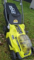 Side/front view of Ryobi cordless lawn mower showing deck, wheels and grass bag