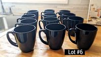 Photo shows 12 black ceramic mugs arranged on a wooden surface in a kitchen setting, demonstrating quantity and condition.